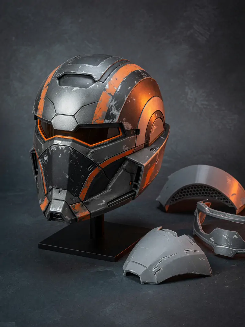 Capacete de cosplay impresso em 3D, pintado em cinza e laranja, ao lado de peças cruas